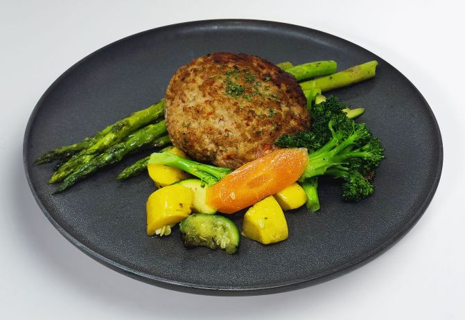 A gourmet turkey burger with grilled asparagus, broccoli, and sliced carrots on a dark plate.