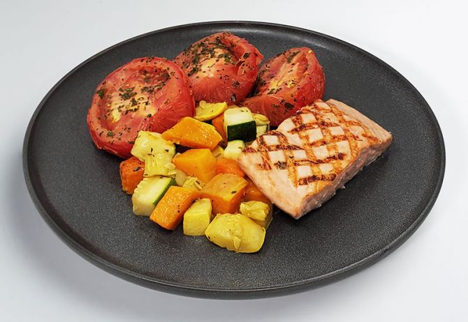 A bowl of grilled salmon, roasted vegetables, and herb baked plum tomato halves on a dark plate.