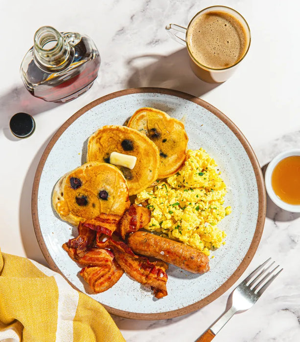 Breakfast plate with pancakes, eggs, bacon, and sausage on a marble surface with coffee and syrup.