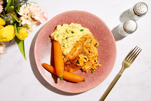 Pink plate with pasta, carrots, and mashed potatoes on a white table with flowers and a fork.