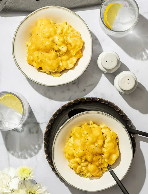 Two bowls of macaroni and cheese with glasses of water and lemon wedges on a white tablecloth.