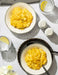 Two bowls of macaroni and cheese with glasses of water and lemon wedges on a white tablecloth.