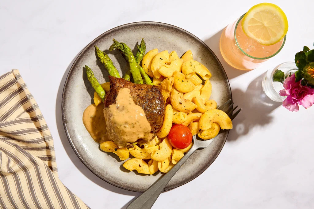 Overhead shot of a plated meal on a white marble surface, featuring a piece of grilled steak topped with creamy sauce, macaroni pasta, green asparagus, and a cherry tomato. A pink drink with a lemon slice and a striped napkin are also visible.