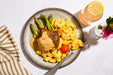 Overhead shot of a plated meal on a white marble surface, featuring a piece of grilled steak topped with creamy sauce, macaroni pasta, green asparagus, and a cherry tomato. A pink drink with a lemon slice and a striped napkin are also visible.
