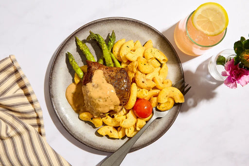 Overhead shot of a plated meal on a white marble surface, featuring a piece of grilled steak topped with creamy sauce, macaroni pasta, green asparagus, and a cherry tomato. A pink drink with a lemon slice and a striped napkin are also visible.