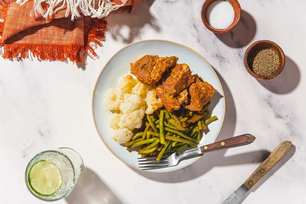 Overhead flat-lay of a plated meal on a white marble surface, featuring breaded chicken pieces, cauliflower, green beans, a fork, a knife, small bowls of seasoning, and a glass with a lime wedge. An orange fringed cloth is visible in the top left.