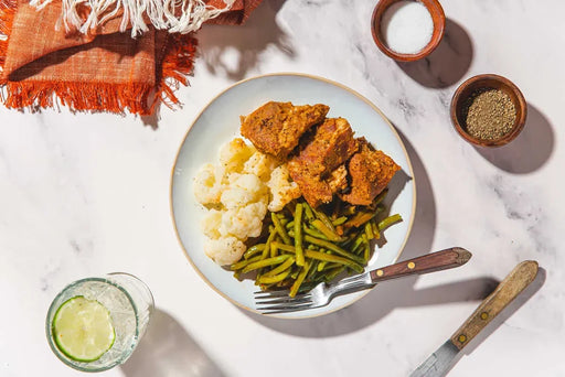 Overhead flat-lay of a plated meal on a white marble surface, featuring breaded chicken pieces, cauliflower, green beans, a fork, a knife, small bowls of seasoning, and a glass with a lime wedge. An orange fringed cloth is visible in the top left.