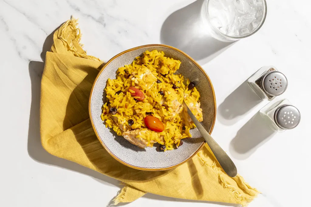 A top-down view of a bowl of yellow rice with beans, black beans, and tomato pieces, placed on a yellow napkin on a white marble surface. A glass of water and salt and pepper shakers are visible in the background.