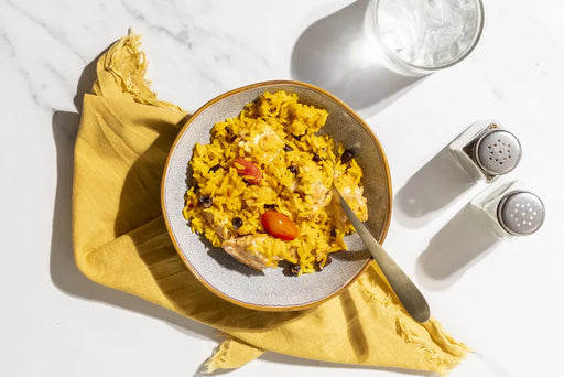 A top-down view of a bowl of yellow rice with beans, black beans, and tomato pieces, placed on a yellow napkin on a white marble surface. A glass of water and salt and pepper shakers are visible in the background.