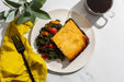 Plate of food with a side of greens and tomatoes, accompanied by a cup of coffee and yellow napkin on a white surface.