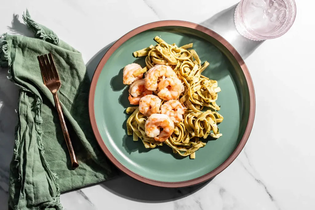 Green plate with shrimp and pasta on a marble surface with a green napkin and fork.