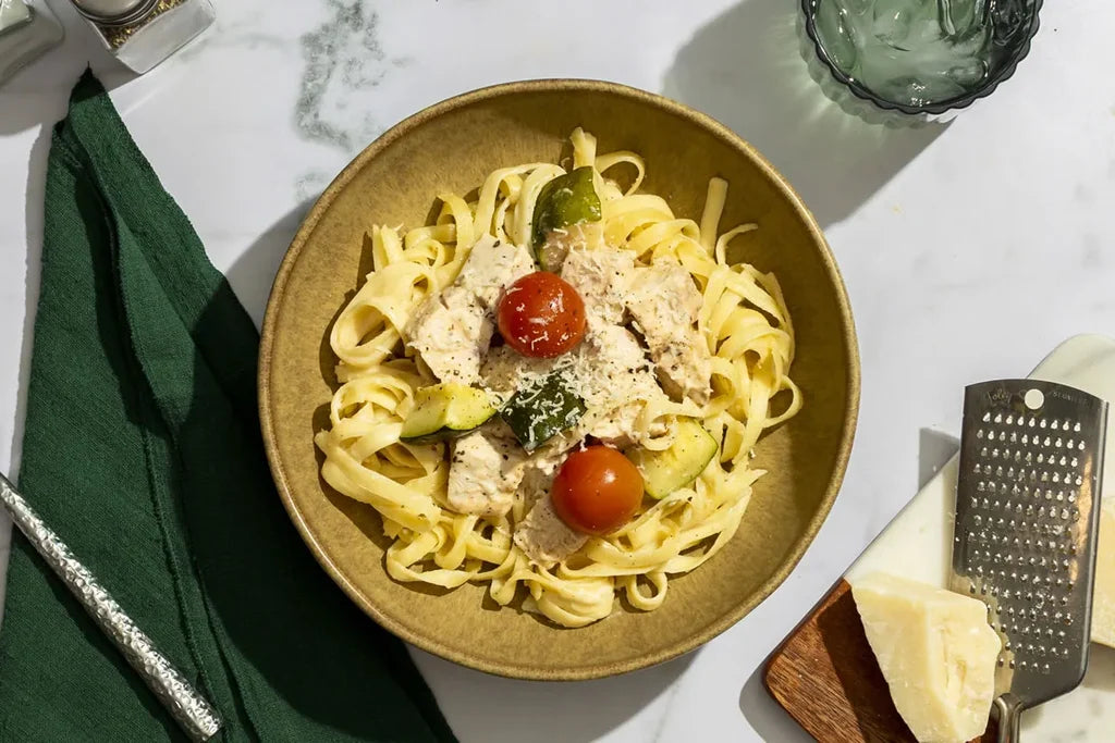 A top-down view of a bowl of pasta with chicken, cherry tomatoes, broccoli, and zucchini on a white marble table, styled with a green napkin, cheese, a grater, and a glass.