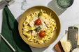 A top-down view of a bowl of pasta with chicken, cherry tomatoes, broccoli, and zucchini on a white marble table, styled with a green napkin, cheese, a grater, and a glass.