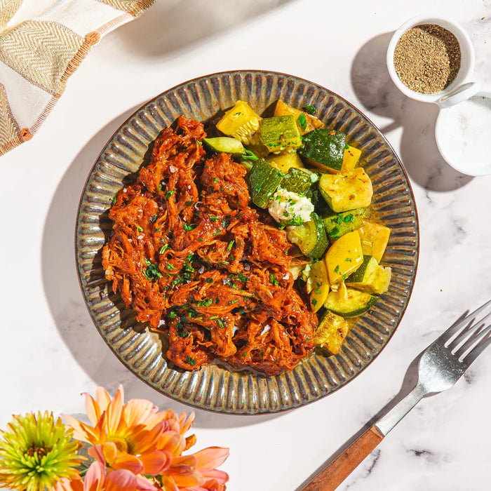 A top-down view of a plated meal on a white marble surface, featuring shredded red meat and cooked yellow and green vegetables on a ribbed gray plate, with a fork, small bowls, and flowers around it.